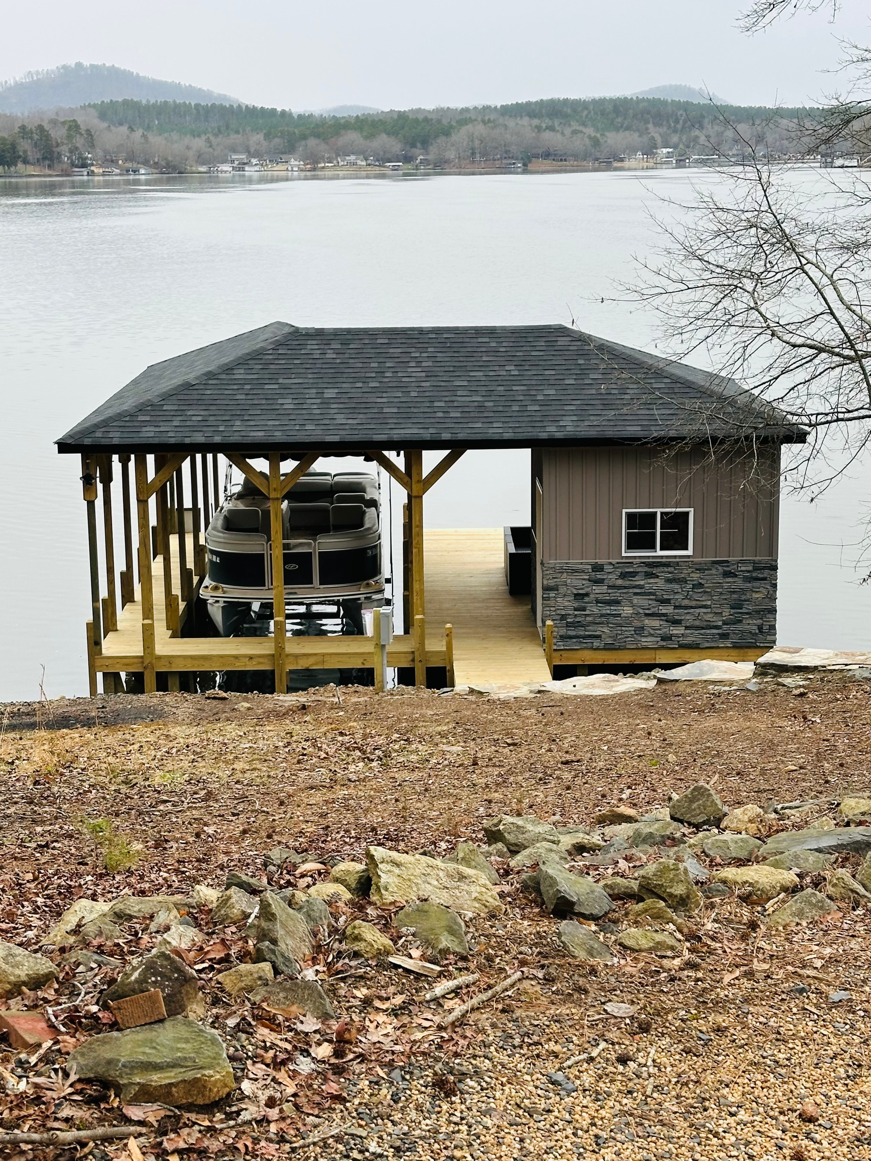 SHT Construction — completed boathouse with dark shingle roof, stone veneer storage room, and pontoon boat on lift