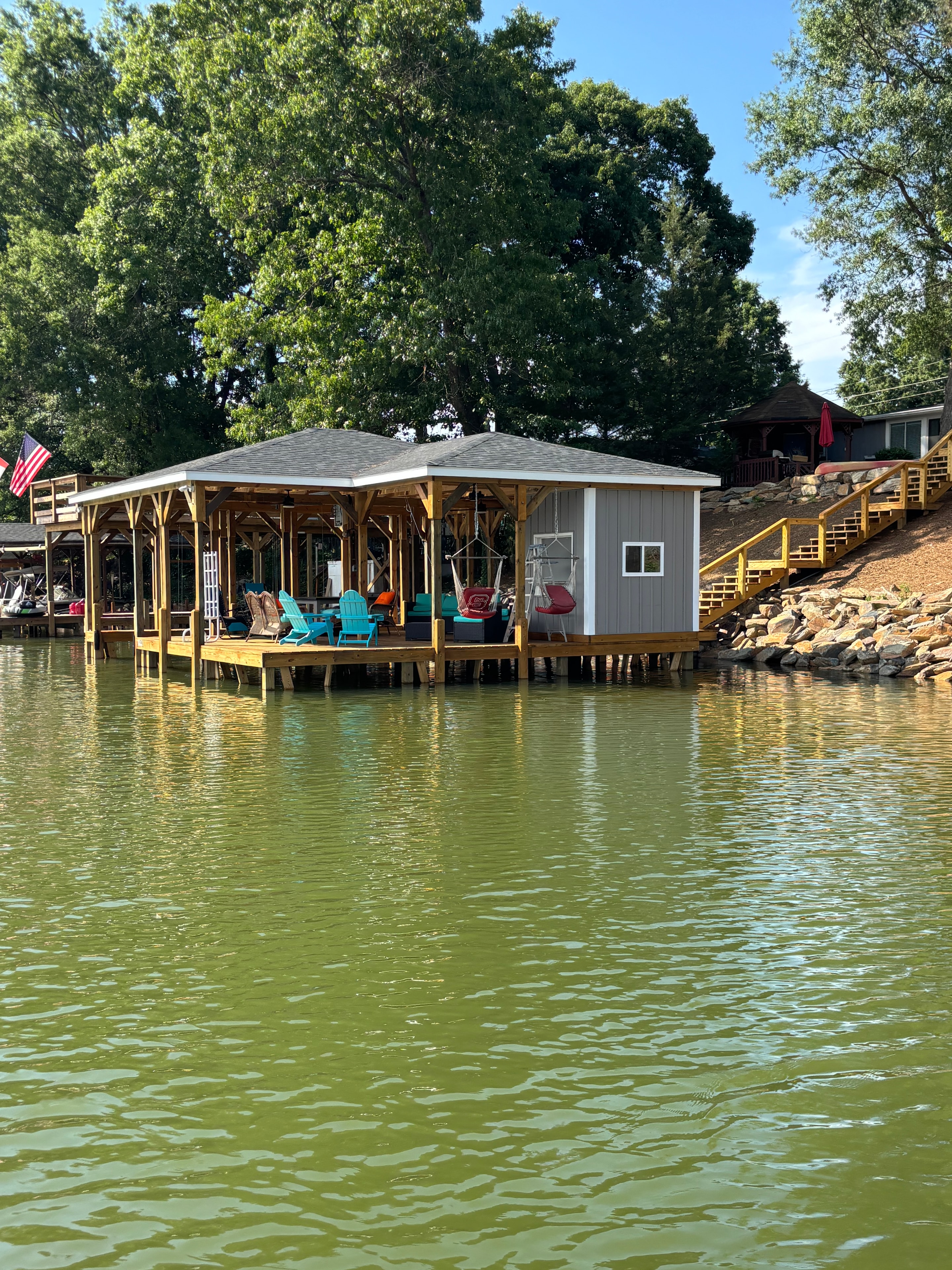 SHT Construction — completed gray boathouse with long dock, colorful Adirondack chairs, and riprap shoreline